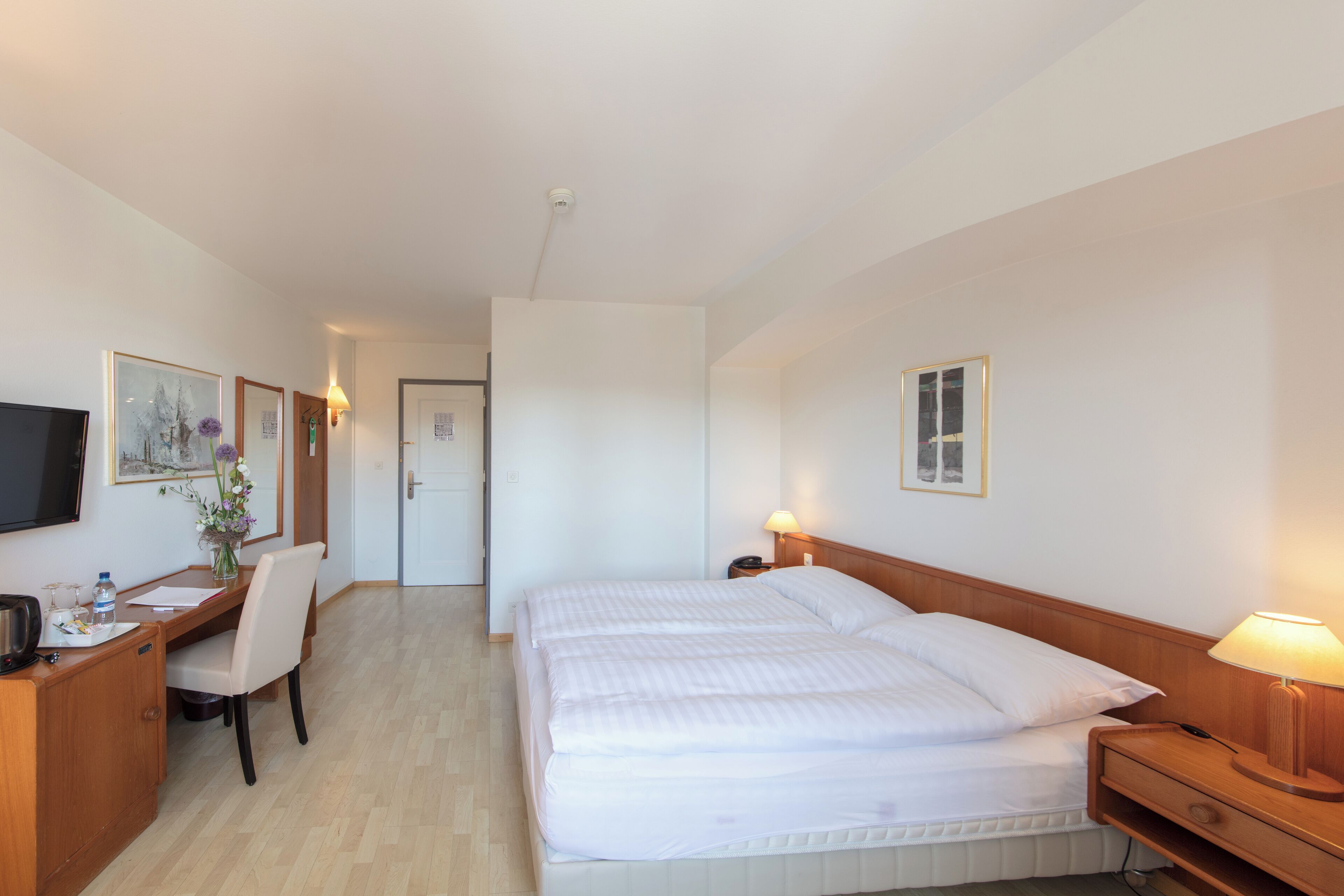 A hotel room with a bed, bedside table, lamp, desk with chair, TV, and artwork on the walls.