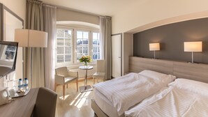 Habitación de hotel con cama, lámparas de noche, un escritorio con lámpara, una silla, una mesita con un florero y una ventana con cortinas.