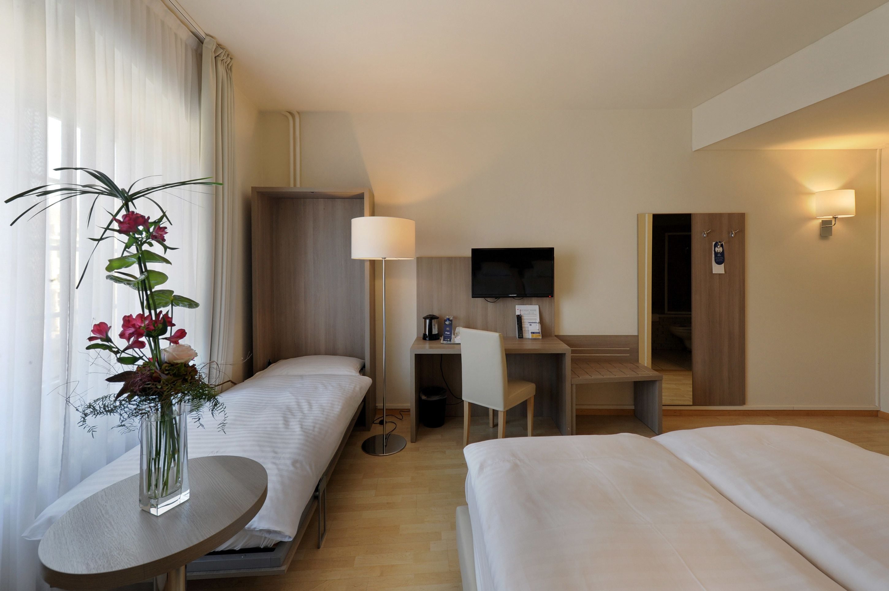 A hotel room with two beds, a desk with a chair, a TV, and a vase with flowers.
