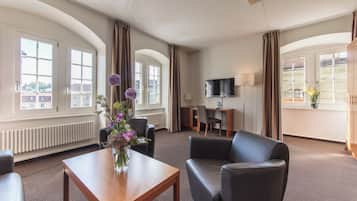 A hotel room with a wooden table, two armchairs, a TV, and a view of the city through large windows.