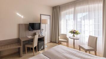 Habitación de hotel con cama, escritorio, silla, televisor y una pequeña mesa con un jarrón de flores.