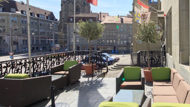 Un espacio al aire libre con mobiliario de mimbre y plantas en macetas, con vista a una calle de la ciudad con edificios históricos y una torre de iglesia.