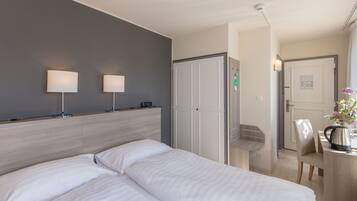 A hotel room with a double bed, two bedside lamps, a wooden headboard, a wardrobe, a desk with a kettle, and a chair.