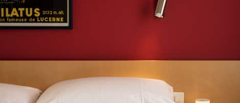 Una habitación de hotel con cama, almohadas blancas, mesita de noche con lámpara y un cartel en la pared.