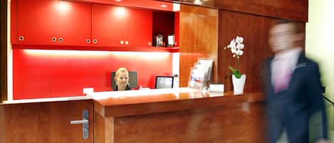 Un mostrador de recepción con detalles de madera y toques rojos, un florero blanco y un monitor de computadora.