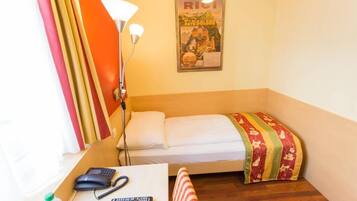 A small hotel room with a single bed, a desk with a phone and remote controls, a lamp, and a poster on the wall.