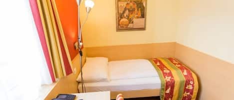 A small hotel room with a single bed, a desk with a phone and remote controls, a lamp, and a poster on the wall.