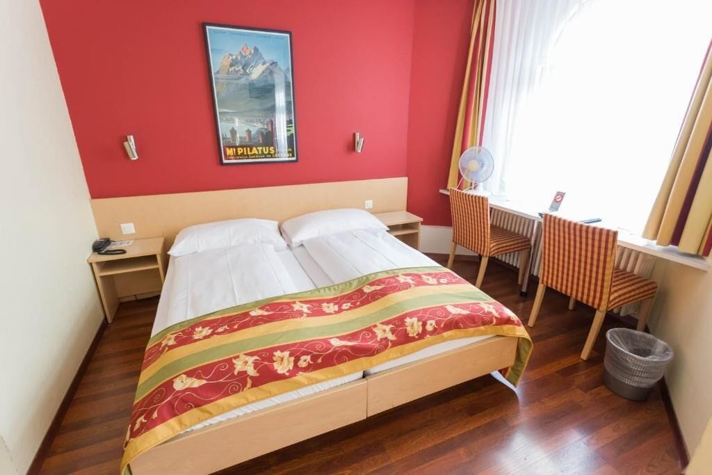 Habitación de hotel con cama, escritorio, silla, un cartel en la pared y ventana con cortinas.