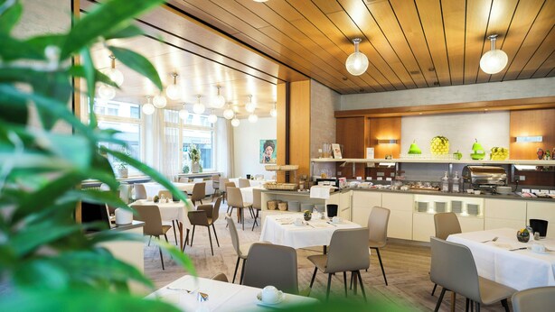 A modern restaurant with round tables, chairs, and a kitchen area. - TOP VCH Hotel Wartburg Stuttgart (Stuttgart)