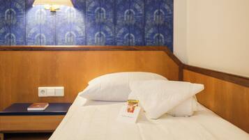 Habitación de hotel con cabecera de madera, cama blanca con almohada, mesita de noche con lámpara y un libro, y pared con diseño.