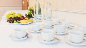 Una mesa dispuesta con tazas y platillos blancos, y una fuente de frutas con manzanas, uvas y plátanos.