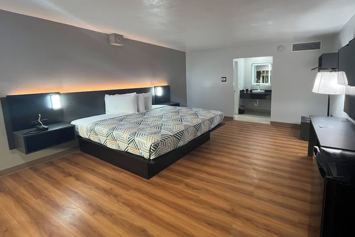 A modern hotel room with a large bed, bedside tables, a desk, a mirror, and a bathroom visible through an open door.