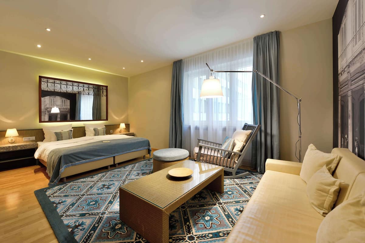 A hotel room with a bed, a sofa, a chair, a table, and a large window with curtains.