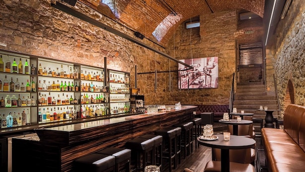 Un bar con pared de ladrillo de fondo, mostrador de bar de madera y varias zonas de estar que incluyen taburetes y mesas redondas.