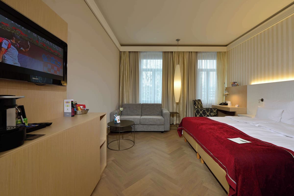 A hotel room with a bed, a sofa, a TV, a coffee machine, and a desk with a lamp.