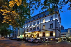 A hotel with a classic facade, a balcony, and a parking area with several cars.