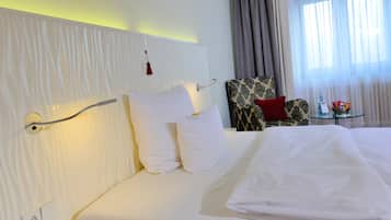 Ein Hotelzimmer mit einem ordentlich bezogenen Bett, einem Stuhl, einem kleinen Tisch mit einer Flasche und Snacks sowie einem Fenster mit Vorhängen.