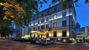 Un hotel con fachada clásica, un acceso bien iluminado y un estacionamiento con varios automóviles.