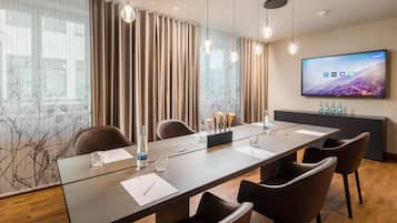 Una sala de conferencias con una larga mesa, sillas, un televisor en la pared y un ventanal con cortinas.