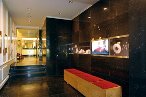 Un hall d’hôtel avec un sol en marbre, un banc en bois doté de coussins rouges et un mur orné d’affiches d’information.