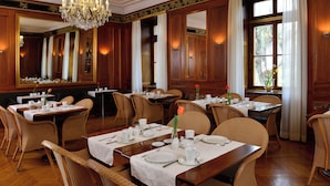 Un comedor formal con paneles de madera, ventanas amplias con cortinas y una araña. Las mesas están dispuestas con manteles blancos y cubiertos.