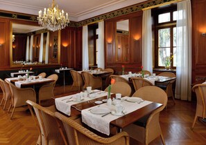 Enjoy a sophisticated dining experience in this elegantly designed room, featuring wooden tables, wicker chairs, and an ornate chandelier.