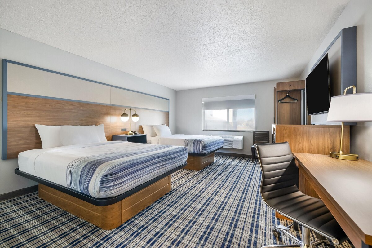 A hotel room with two beds, a desk, a chair, a TV, and a window.