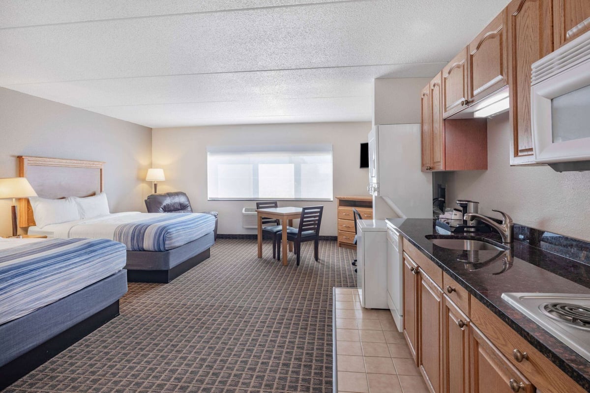 A compact hotel room with a bed, a small dining table, a kitchenette, and a window.