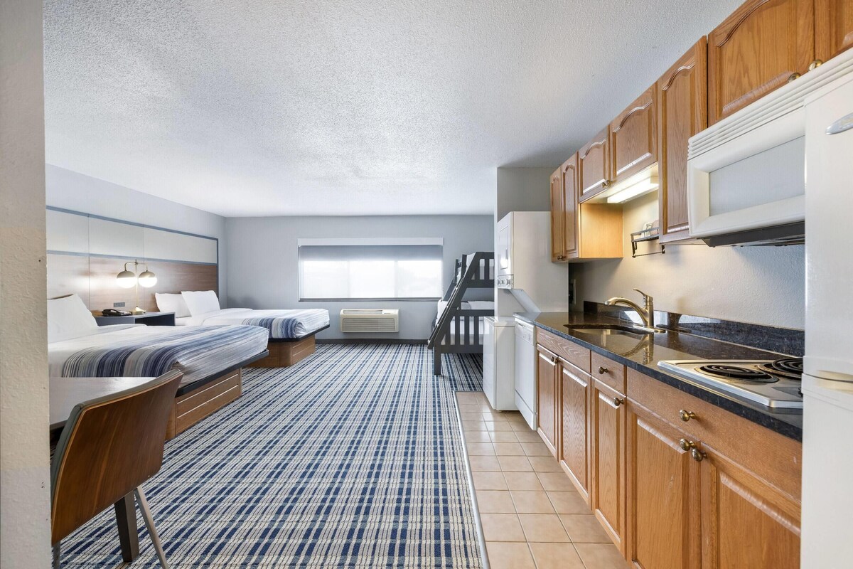 A compact hotel room with a kitchenette, two beds, a large window, and a patterned rug.