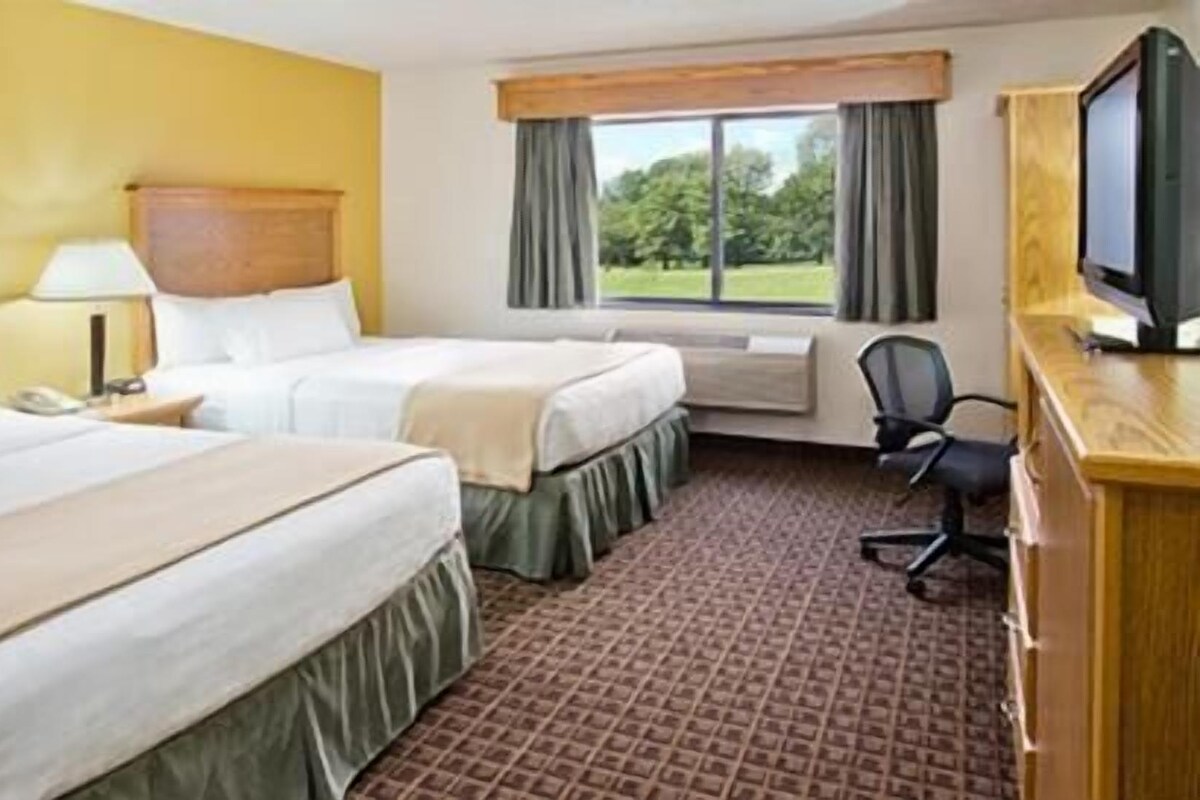 A hotel room with two beds, a desk with a computer, an air conditioning unit, and a window with curtains.