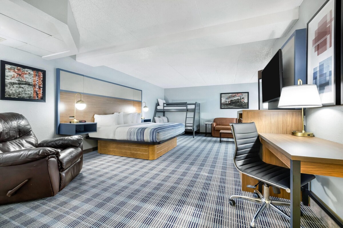 A hotel room with a bed, a desk, a chair, a sofa, a TV, and artwork on the walls.