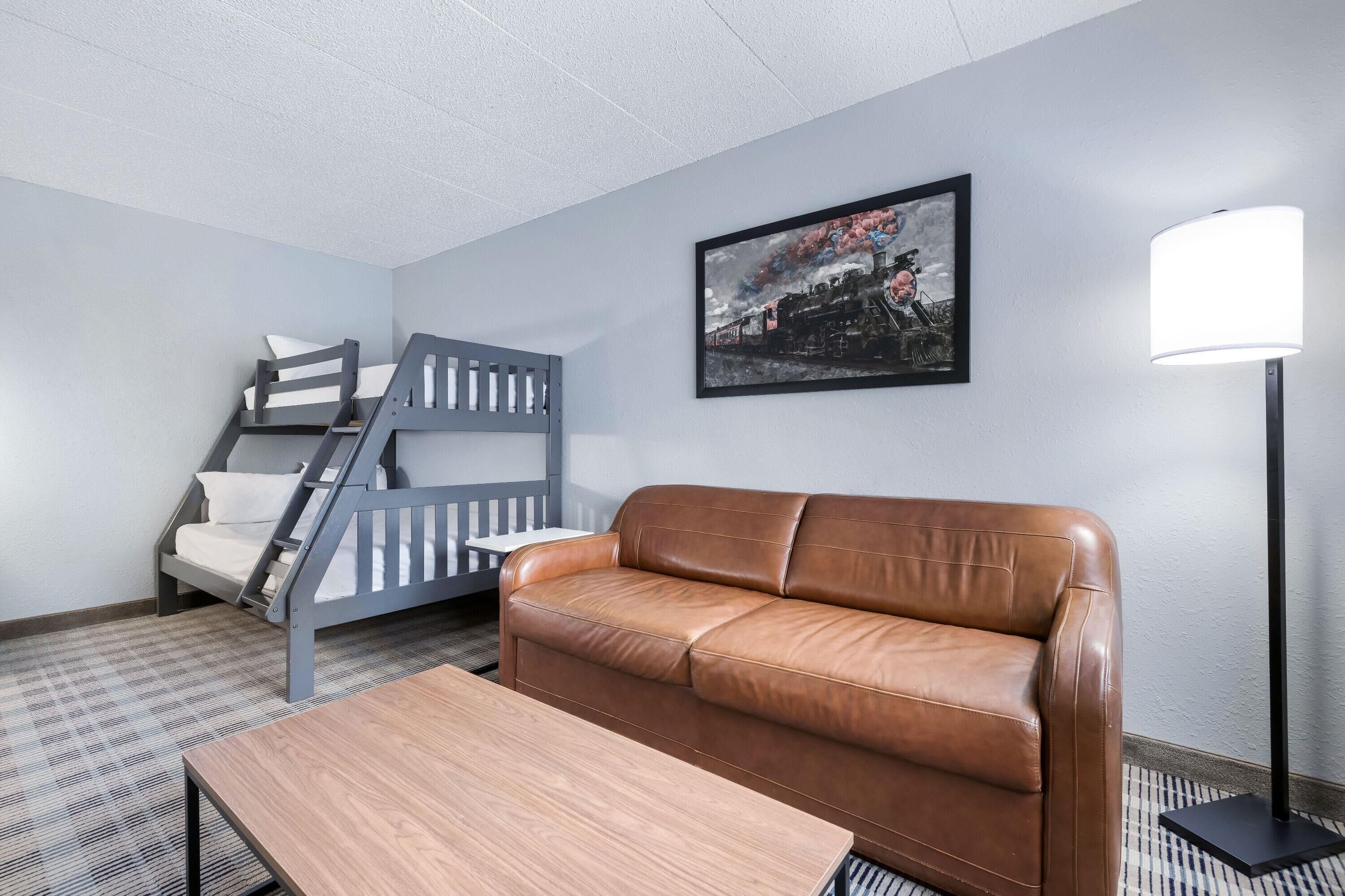 A room with a bunk bed, a leather sofa, a wooden table, a floor lamp, and a framed picture on the wall.
