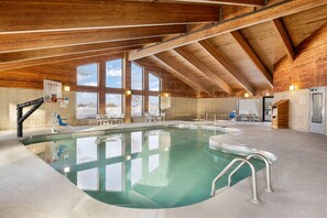 An indoor swimming pool with a wooden ceiling, clear glass walls, and a basketball hoop.