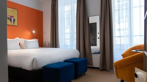 A hotel room with a large bed, two blue ottomans, a yellow armchair, a mirror, and a poster on the wall. - ClapClap Hôtel Strasbourg (Strasbourg)