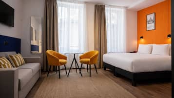 A hotel room with a bed, a sofa, two chairs, a small table, a mirror, and a wall-mounted TV.