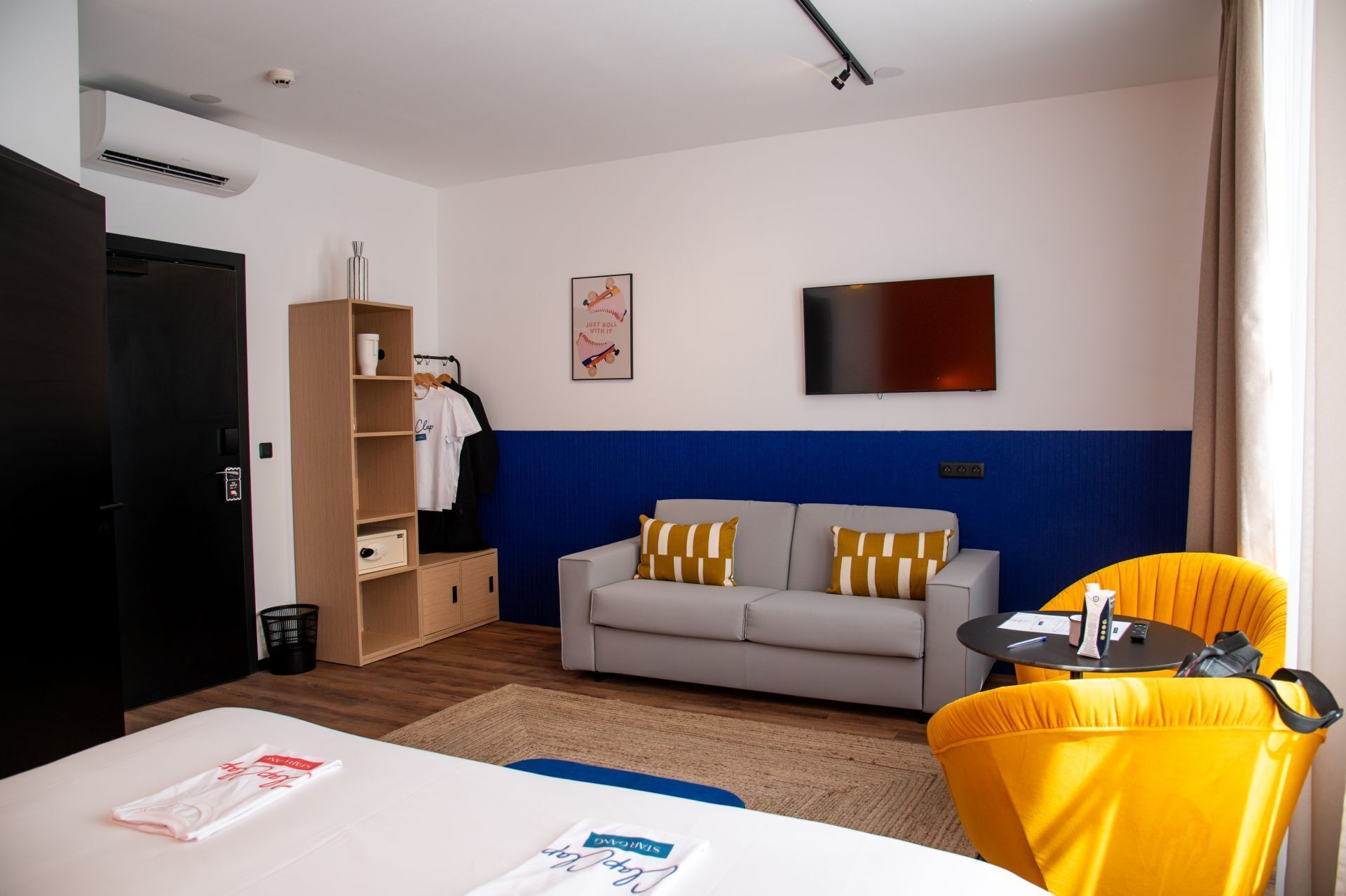 A hotel room with a bed, a sofa, a small table, a chair, a wardrobe, a TV, and a wall-mounted air conditioning unit.
