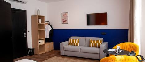 A hotel room with a bed, a sofa, a small table, a chair, a wardrobe, a TV, and a wall-mounted air conditioning unit.