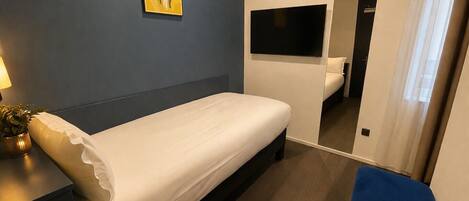 Una habitación de hotel con una cama, un televisor montado en la pared, una mesita de noche con lámpara y una planta en maceta.