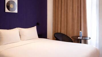 A hotel room with a large bed, a small table, a chair, a wall-mounted light fixture, and a window with curtains.