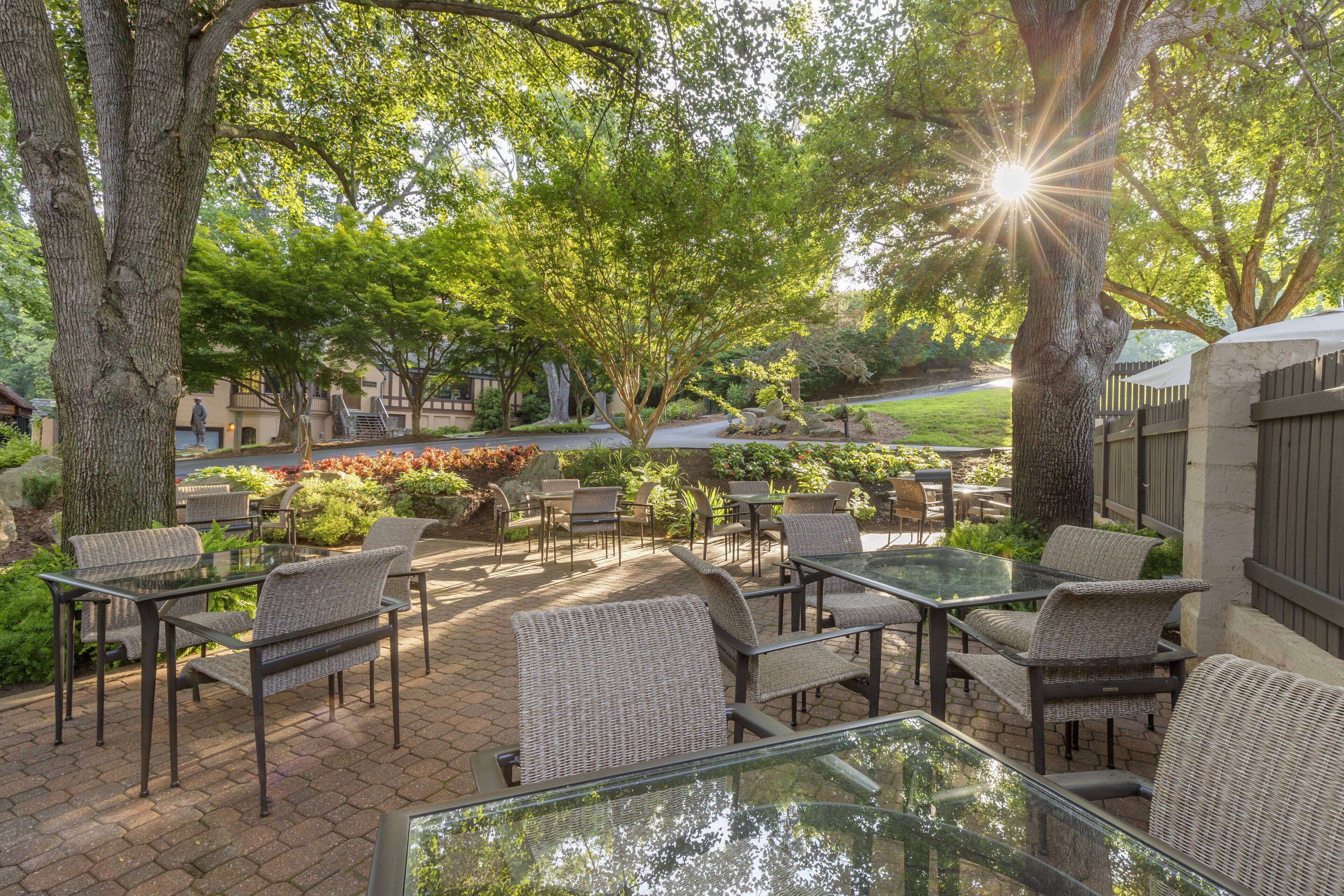 An outdoor patio with wicker furniture and glass tables, surrounded by trees and a view of a residential area.