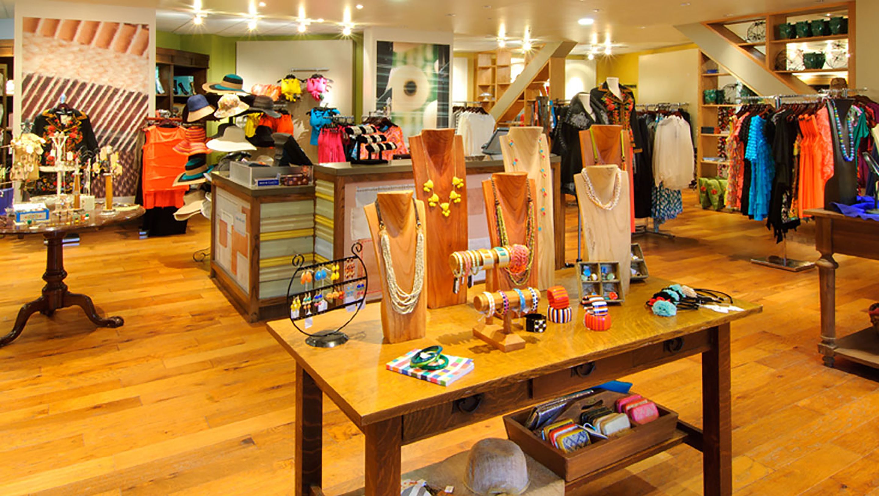 Una boutique con piso de madera, una mesa de exhibición central con joyería y accesorios, y percheros de ropa al fondo.