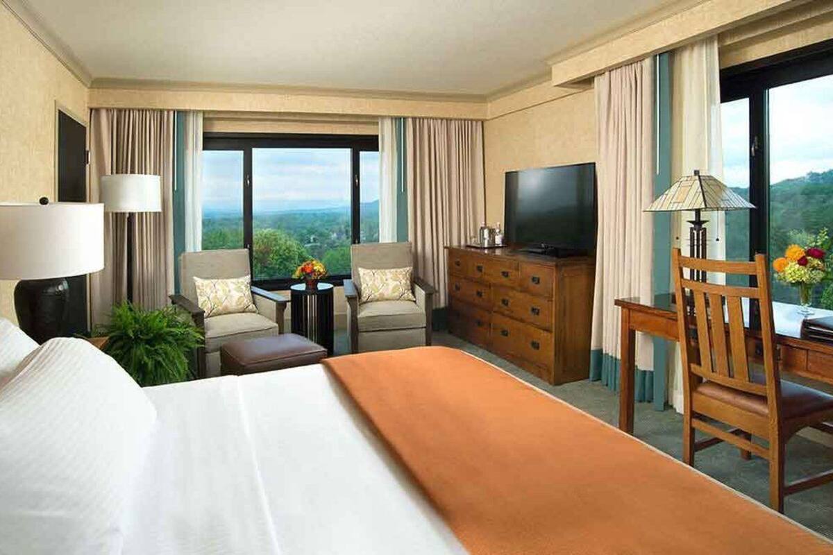 A hotel room with a large bed, a desk with a chair, a television, a dresser, and a view of the outdoors.