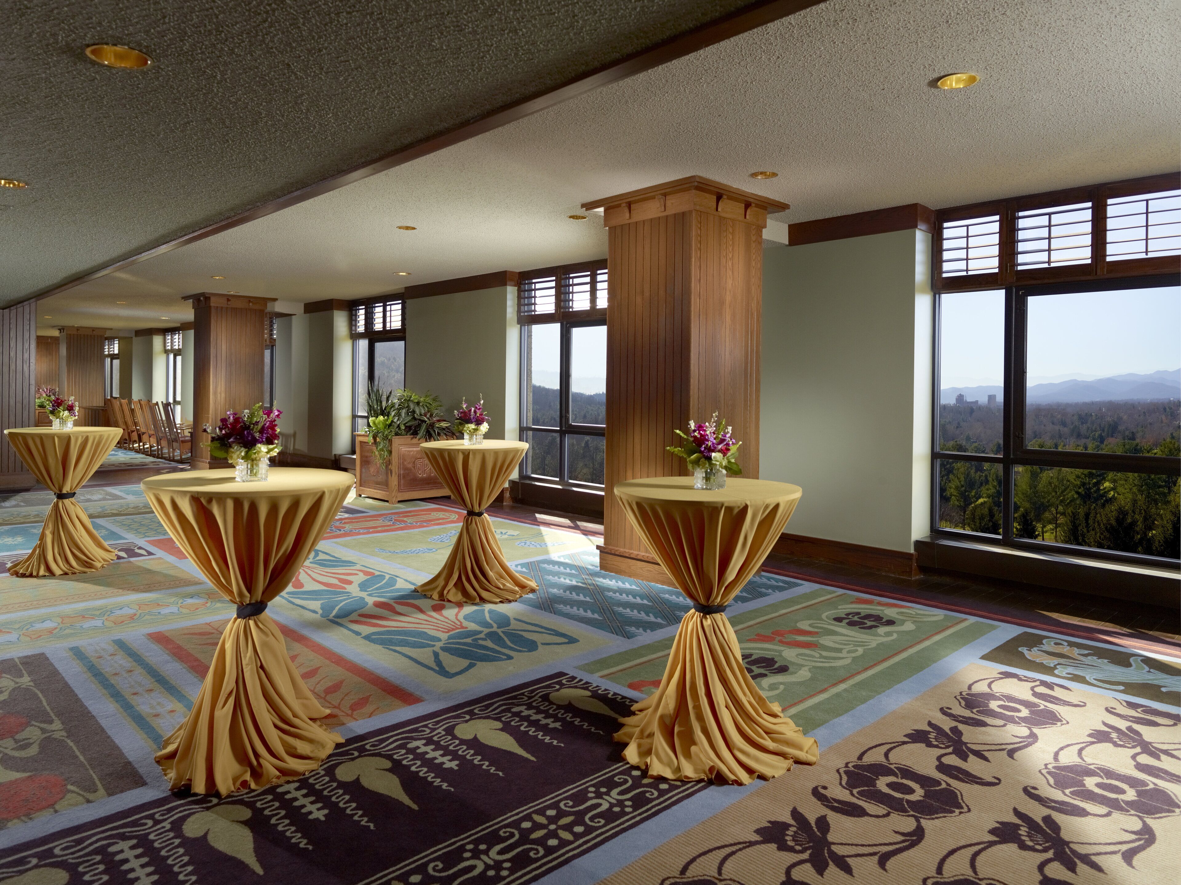 Un amplio vestíbulo de hotel con techos altos, grandes ventanales que ofrecen vistas a las montañas y múltiples mesas redondas con manteles cubiertos y centros de mesa florales.