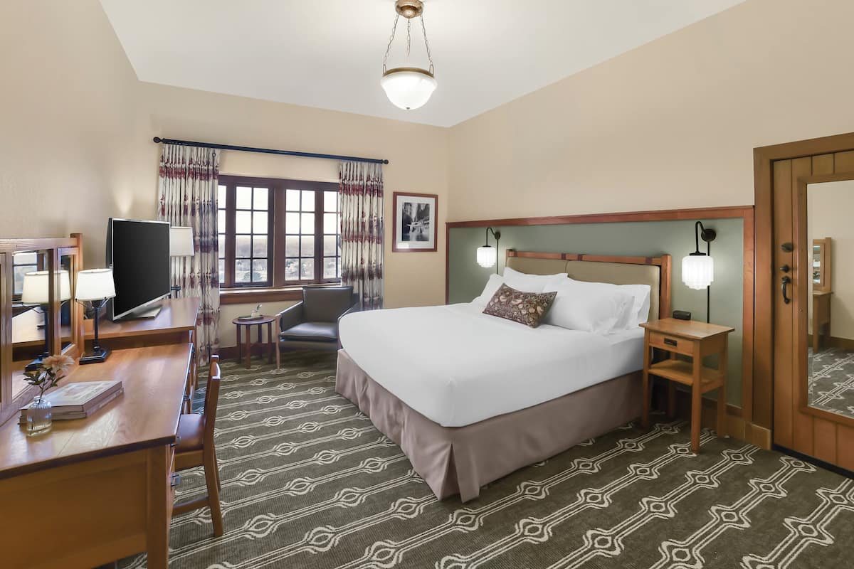 A hotel room with a large bed, a desk with a computer, a chair, a small table, and a window with curtains.