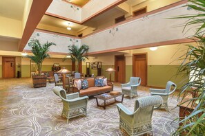 A spacious lobby with a central seating area, multiple chairs, a sofa, and a coffee table. There are two large potted plants and a wooden cabinet. The floor has a patterned carpet.