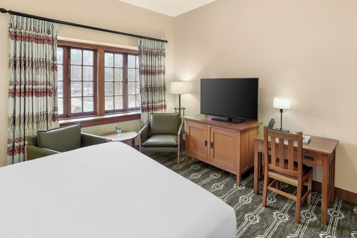 A hotel room with a bed, a desk with a television, a chair, a small table, and a window with curtains.