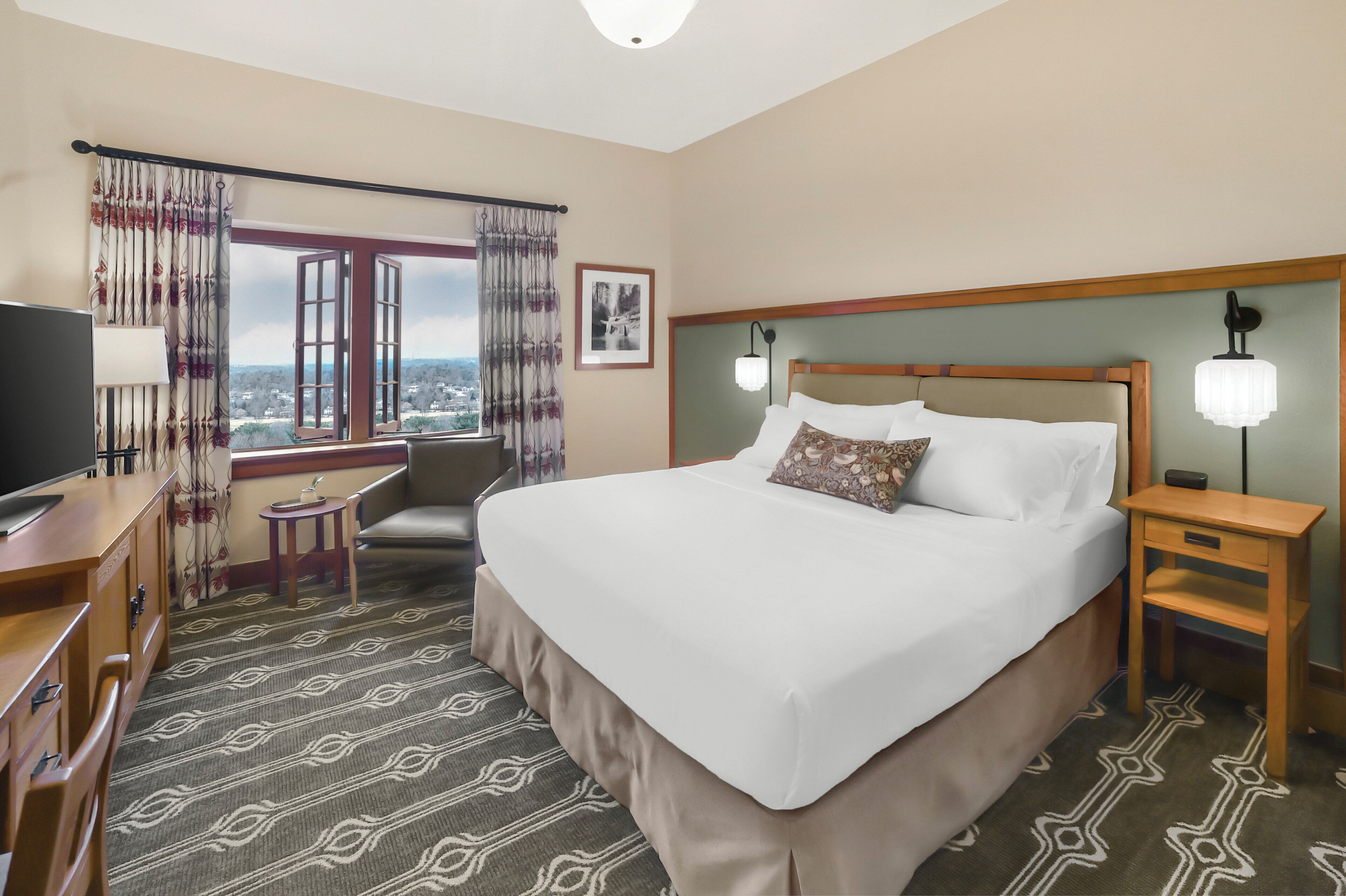 A hotel room with a large bed, a desk, a chair, a small table, and a window with curtains.