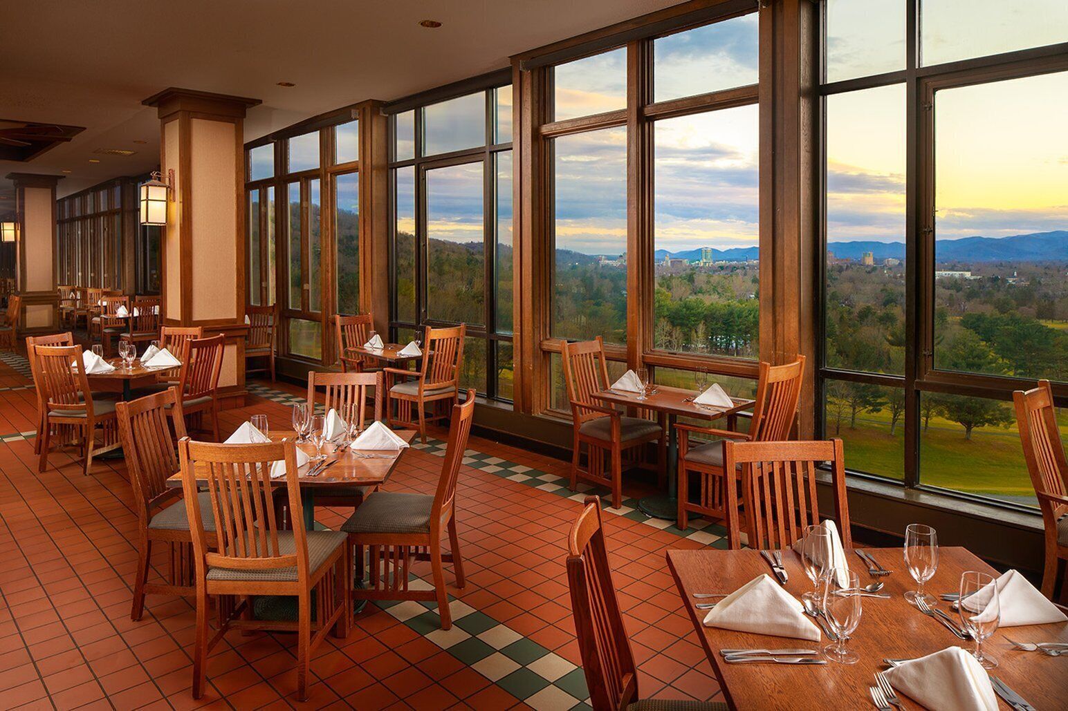 Un restaurante con amplios ventanales que ofrece una vista panorámica de un paisaje montañoso. Las mesas están preparadas con servilletas blancas y cubiertos.