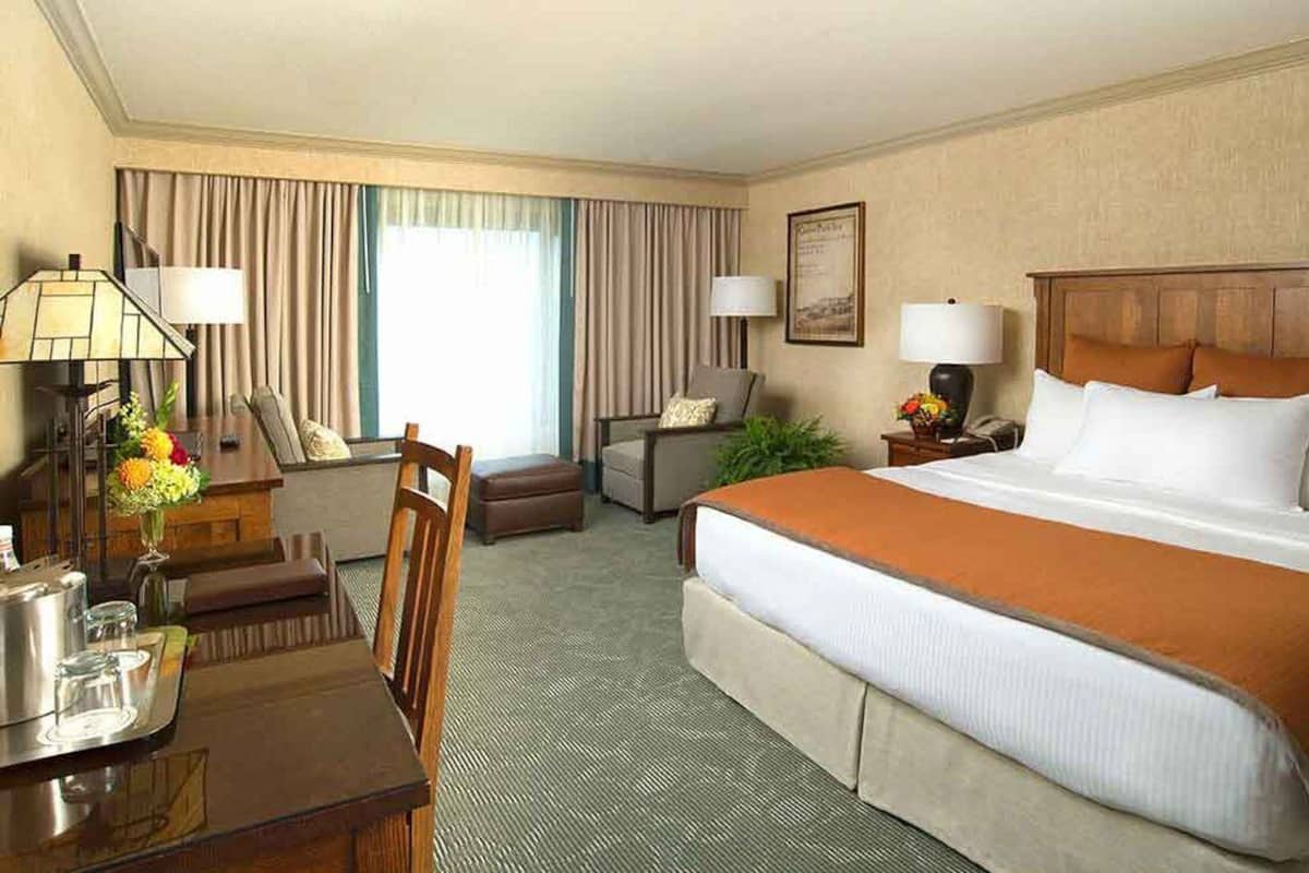 A hotel room with a large bed, a desk with a lamp, a chair, a small table with a tray, and a window with curtains.