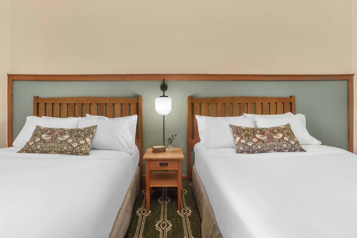 A hotel room with two beds, each with white bedding and floral-patterned pillows. There is a nightstand with a lamp between the beds.